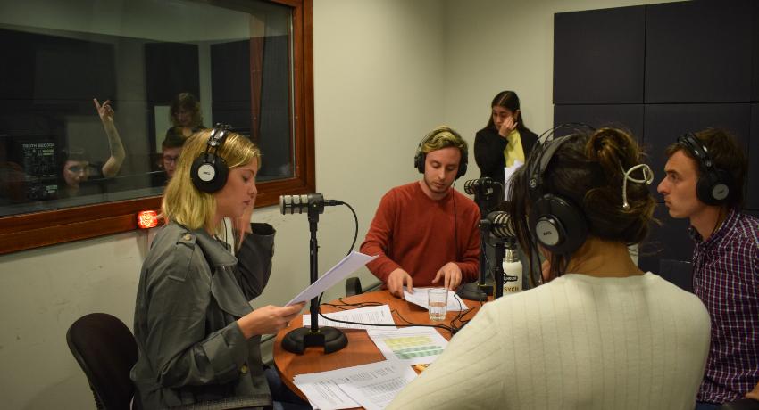 Cinco estudiantes en un estudio de radio, con auriculares en torno a una mesa. Se ve una ventana y del otro lado el operador levanta la mano para indicar algo. 
