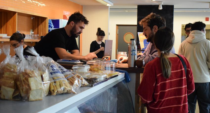 Fotografía de la cantina de la FIC: en primer plano sobre el mostrador se ven paquetes con grisines y galletas, y más atrás una mujer con buzo a rayas rojo y rosado, con el pelo atado y un hombre castaño sosteniendo una tarjeta de débito. Del otro lado del mostrador orto hombre morocho, alto de remera negra y barba digita algo en un pos.