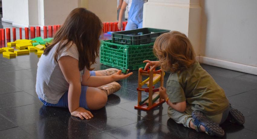 Foto de un niño y una niña de espaldas juegan con maderas sentados en el piso del hall del portal rabú. 