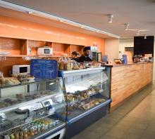 Fotografía de la cantina. Se ven dos vitrinas con productos de panadería adentro y encima y a continuación un mostrador de madera. Sobre una de las vitrinas hay un cartel con una lista de precios, que dice, en fondo azul con letras blancas, Combos de café. Detrás del mostrador hay dos personas, un hombre alto morocho de barba candado y una mujer morocha de pelo corto atado. Visten remeras negras. Se ven dos microondas y una heladera. 